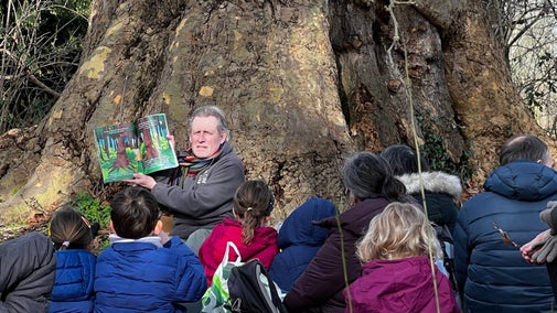 A storyteller sits at the base of a huge, gnarled tree trunk, holding open an illustrated picture book while reading to a group of children and adults seated on the ground. The audience, wearing colourful outdoor coats, gathers closely in a woodland setting lit by winter sunlight.